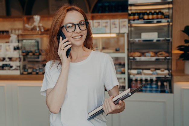 Happy beautiful businesswoman talking on cell phone outside, standing with laptop near coffee shop outdoors, cheerful female freelancer making phone call and smiling while walking in morning city