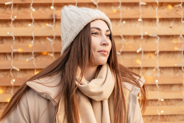 Happy beautiful dreamy girl in trendy winter clothes with a knitted hat, fashion jacket and scarf near a wooden wall with lights