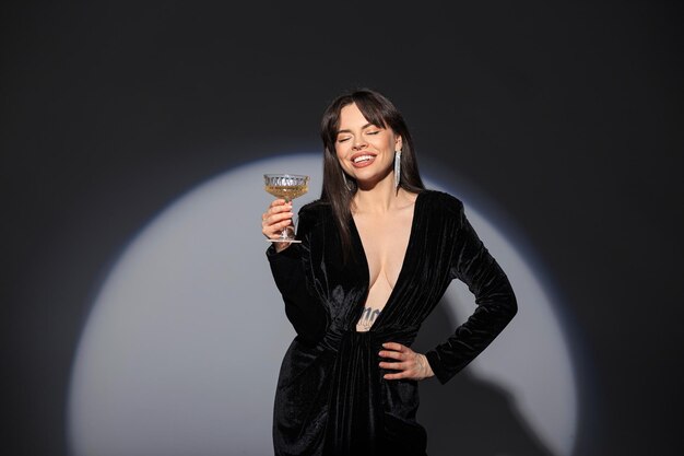 Happy beautiful elegant woman model with a sweet smile in a black sexy fashion dress with a neckline holds a glass of champagne and celebrates in the studio Pretty chic lady