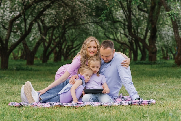 Happy beautiful family is sitting on a blanket in the park in the summer and looking at the blanket talking via video link