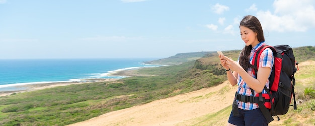 happy beautiful female hiker using the mobile smart phone viewing hiking information through online map guidebook on the top of coastal landscape with banner crop for copy space.