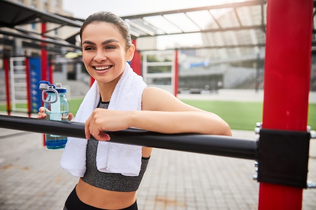 Happy beautiful female is standing near iron bars outdoors with towel and bottle of water