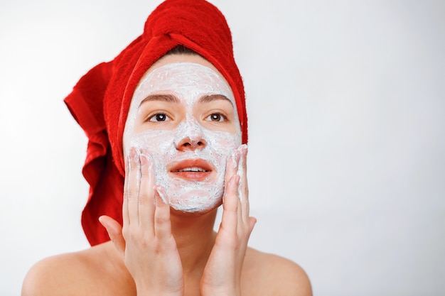 Happy beautiful girl with a red towel on her head applies a scrub on the face of a large portrait on a white background