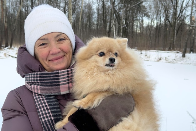 Happy beautiful lady retired elderly senior woman is walking with her Pomeranian Spitz dog at winter cold day in snow