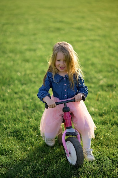 A happy beautiful little girl runs fast and rides her first bicycle without pedals on the grass on a warm summer day Cute baby 3 years old Side view Learning to keep balance