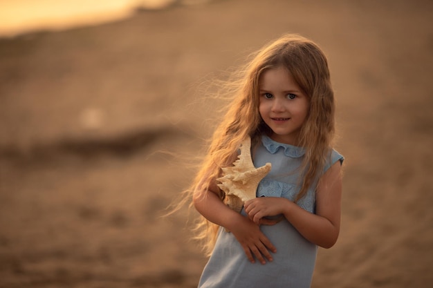 Happy beautiful little girl with long hair on a sandy beach with a starfish in her hands smiling in the rays of the sunset Vacation concept at sea traveling with children camp relaxation spa