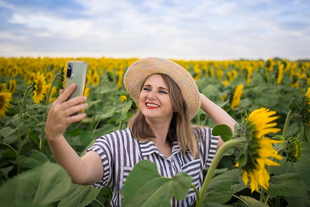 Happy beautiful middle aged woman in a straw hat takes a selfie shoot and rejoices and enjoys the flowering of a sunflower field on a sunny day