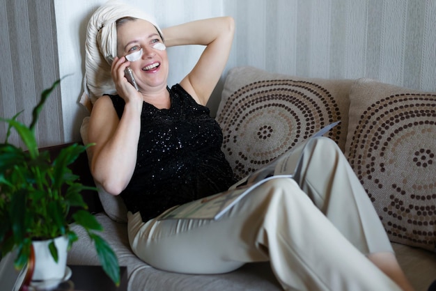 Happy beautiful middle aged woman talking on the phone at home on the sofa after a shower Self care concept Senior's active life position Modern technologies