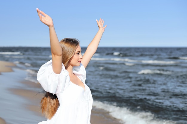 Happy, beautiful woman on the ocean beach standing in a white summer dress, raising hands.