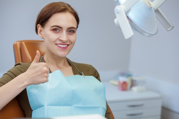 Happy beautiful woman sitting in dental chair, smiling showing thumbs up