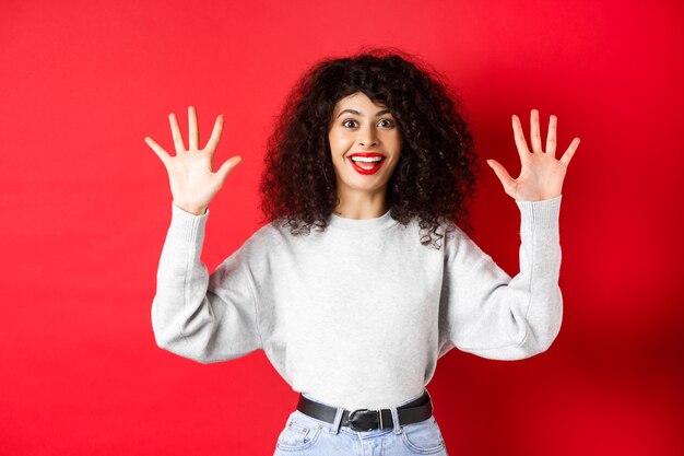 Happy beautiful woman smiling and raising hands up, showing number ten, ordern dozen of something, standing in sweatshirt on red background.