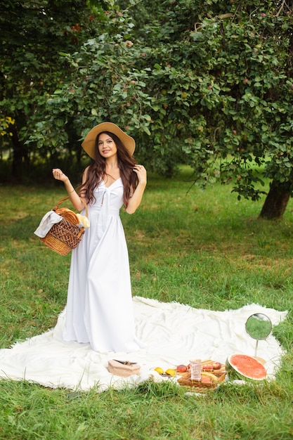 Happy beautiful woman wearing brimmed hat and white dress while standing and holding basket of bread in at summer park