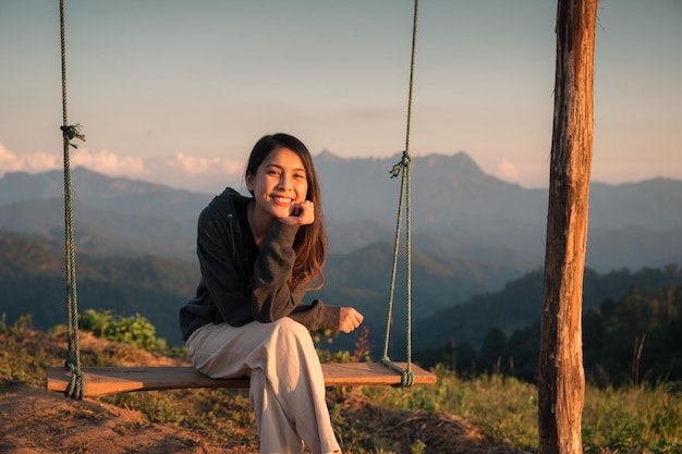 Happy beautiful young asian woman sitting and smiling at wooden swing on top of mountain in countryside at the evening