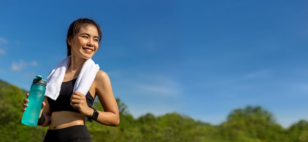 Happy beautiful young Asian woman with her white towel over her neck, standing smiling and holding her water bottle to drink after her morning exercise at an outdoor park