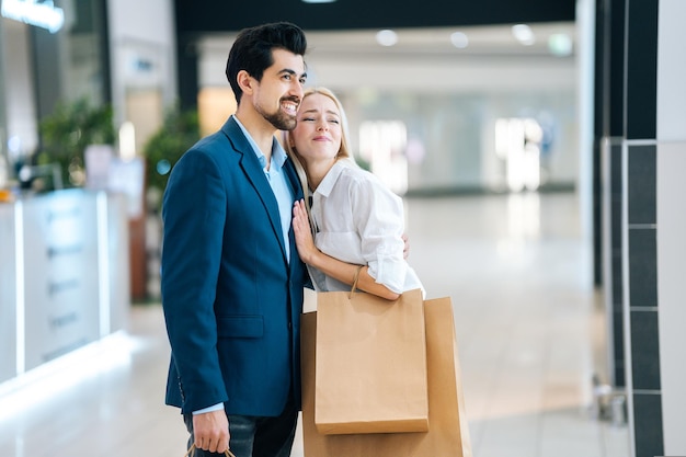 Happy beautiful young couple holding shopping paper bags with purchases and hugging looking upon