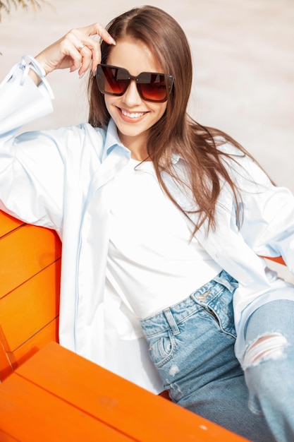 Happy beautiful young fashion woman with a smile with vintage sunglasses in fashionable casual clothes with a blue shirt tshirt and jeans sits and enjoy on a sunny day