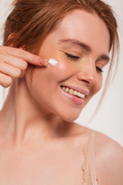 Happy beautiful young fresh redheaded woman model with a cute smile applies cream to face skin and has fun on a white background Beauty and skin care