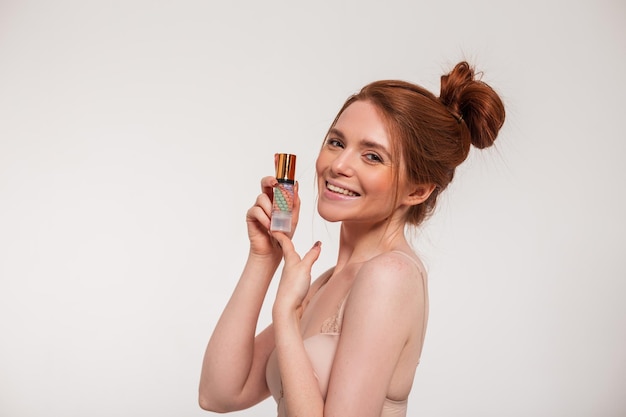 Happy beautiful young fresh woman model with red hair and cute smile holding makeup base cream and having fun on white background Beauty and makeup