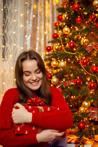 Happy beautiful young girl in red sweater with brown hair hugs a big present box Christmas tree and garland lights on the background