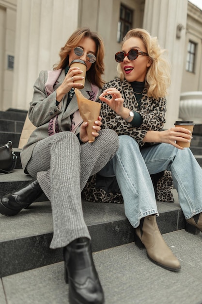 Happy beautiful young girls in fashionable clothes sit on the steps drinking coffee and eating sweets in the city Travel concept