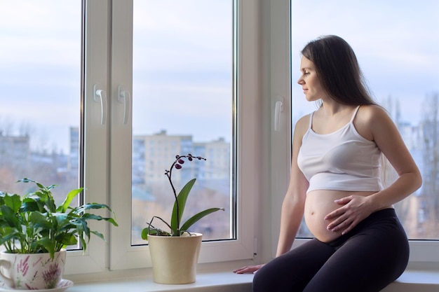 Happy beautiful young pregnant woman dreamily looking out home window