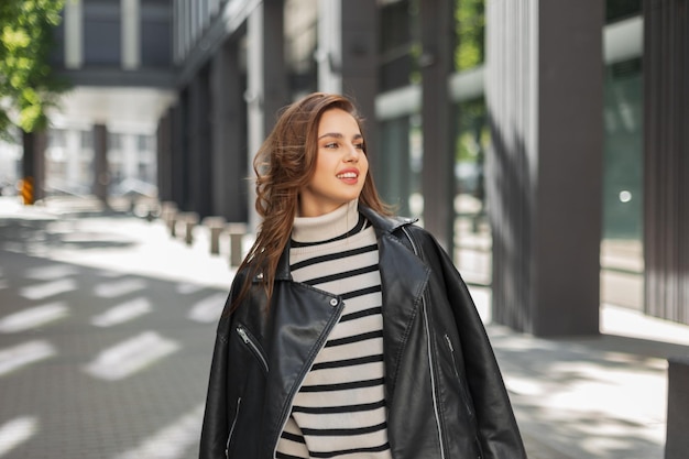 Happy beautiful young stylish girl with a smile in fashionable casual clothes with a leather jacket and a striped sweater walks in the city