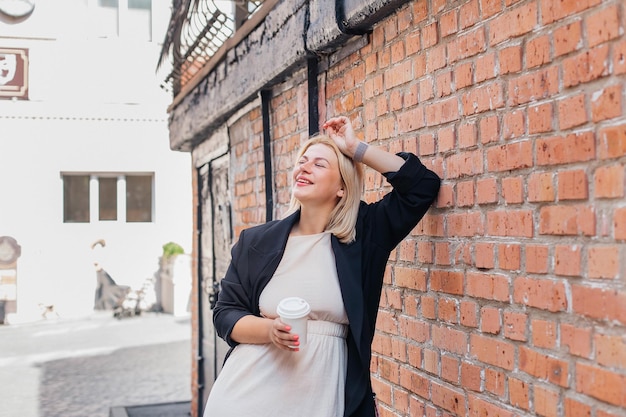 Happy beautiful young woman in a dress and jacket stands by a brick wall and drinks coffee