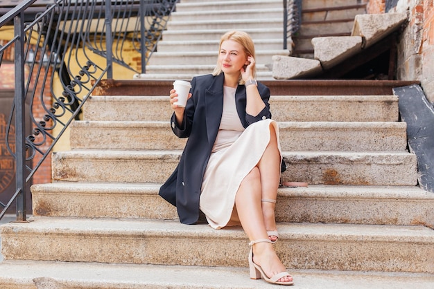 Happy beautiful young woman in dress and jacket walks around the city and drinks coffee