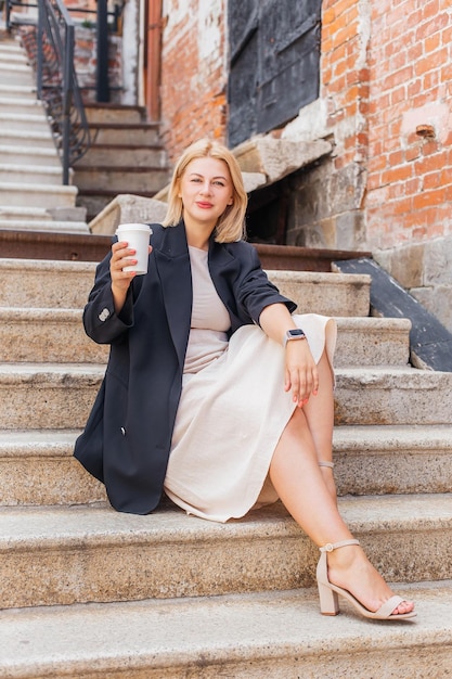 Happy beautiful young woman in dress and jacket walks around the city and drinks coffee