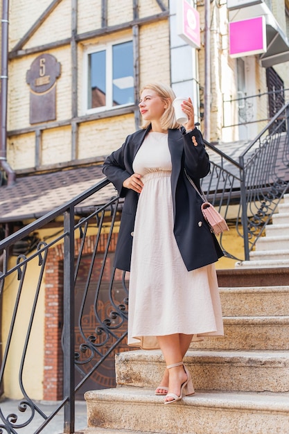 Happy beautiful young woman in dress and jacket walks around the city and drinks coffee