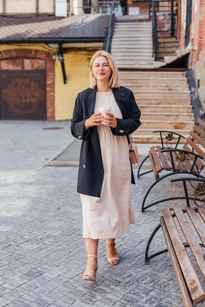 Happy beautiful young woman in dress and jacket walks around the city and drinks coffee