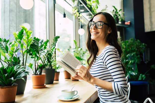 Happy beautiful young woman in glasses is reading book and drinking coffee in modern cafe