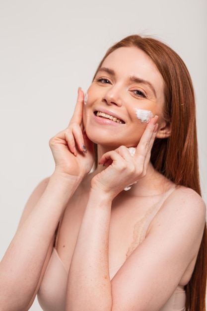 Happy beautiful young woman model with natural red hair and cute smile applies cream on face skin in studio on white background Beauty and skin care