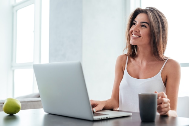 Happy beautiful young woman using laptop and drinking coffee on the kitchen