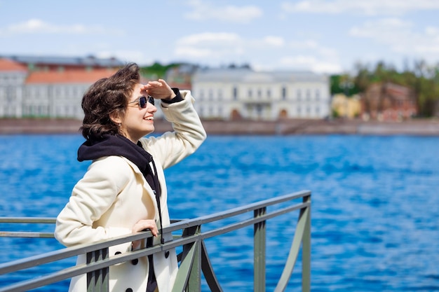 Happy beautiful young woman in white coat and sunglasses standing on embankment