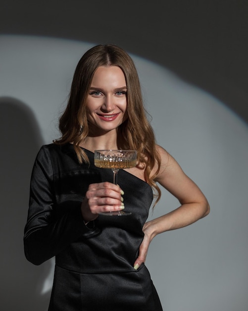 Happy beautiful young woman with a smile in a fashionable black dress holds a vintage glass of champagne on a dark background with circular light Lady celebrates an event