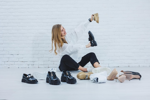 Happy beautiful young woman with a smile in a fashionable outfit with a white shirt and black trousers sits on the floor among the shoes and chooses a new pair of shoes for herself