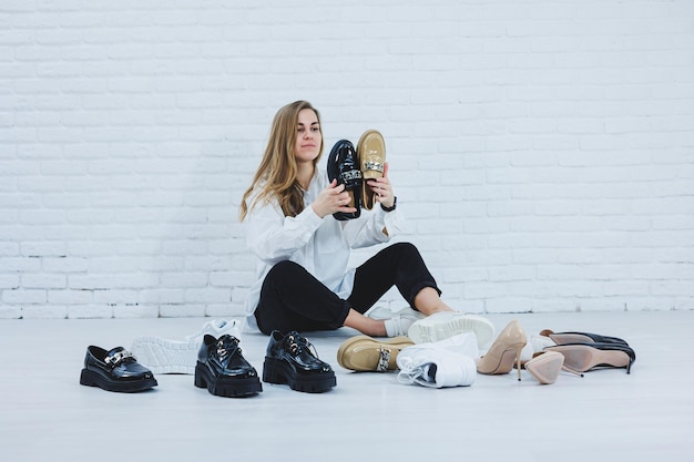 Happy beautiful young woman with a smile in a fashionable outfit with a white shirt and black trousers sits on the floor among the shoes and chooses a new pair of shoes for herself