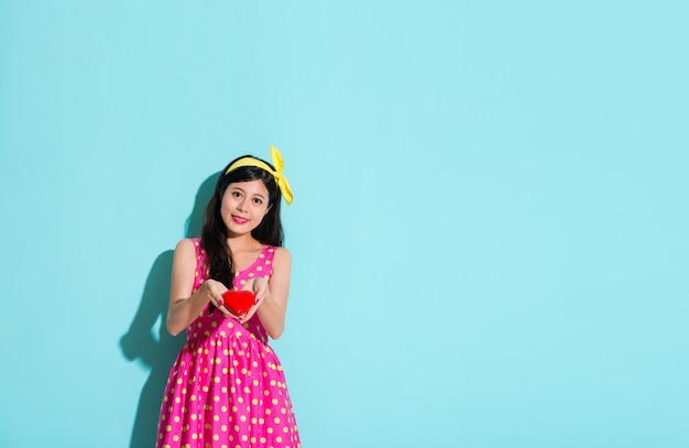 happy beauty lady presenting heart shaped model standing in blue background and wearing cute dress looking at camera smiling.