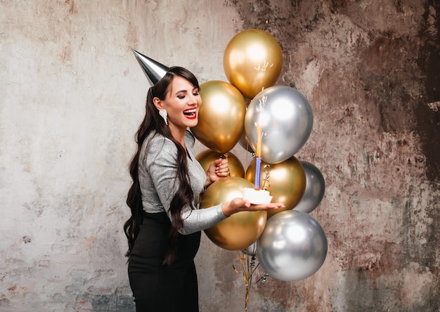 Happy Birthday Sexy brunette posing with balloons fireworks birthday cake on the background of a decorative wall a woman blows out the candles on the cake