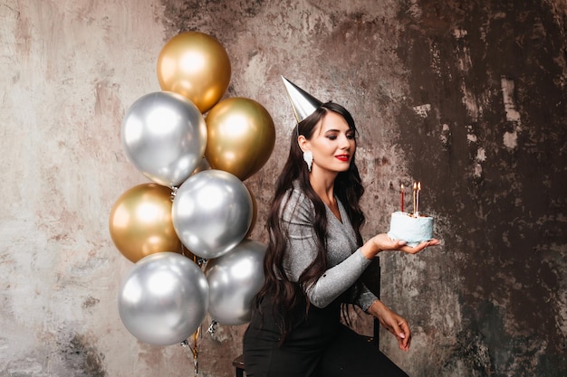 Happy Birthday Sexy brunette posing with balloons fireworks birthday cake on the background of a decorative wall a woman blows out the candles on the cake