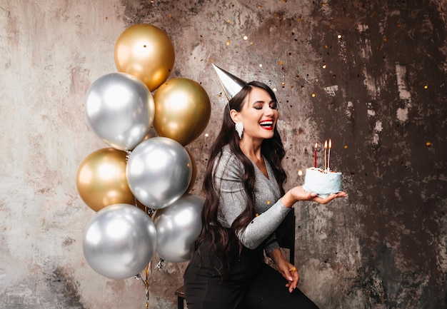 Happy Birthday Sexy brunette posing with balloons fireworks birthday cake on the background of a decorative wall a woman blows out the candles on the cake