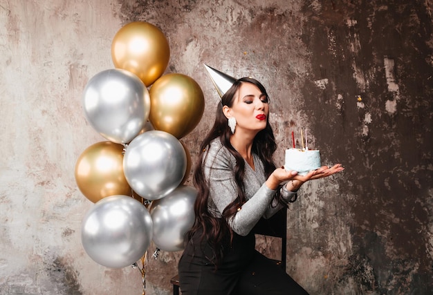 Happy Birthday Sexy brunette posing with balloons fireworks birthday cake on the background of a decorative wall a woman blows out the candles on the cake