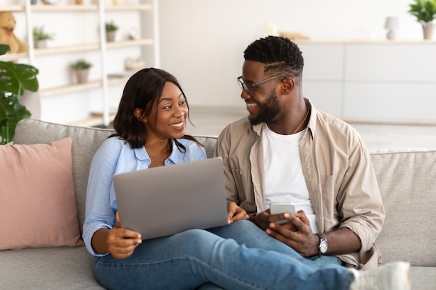 Happy black couple sitting on couch using cellphone and pc