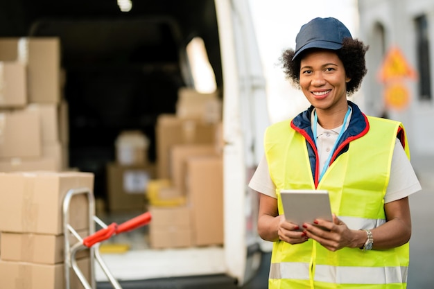 Happy black courier with touchpad delivering packages and looking at the camera outdoors