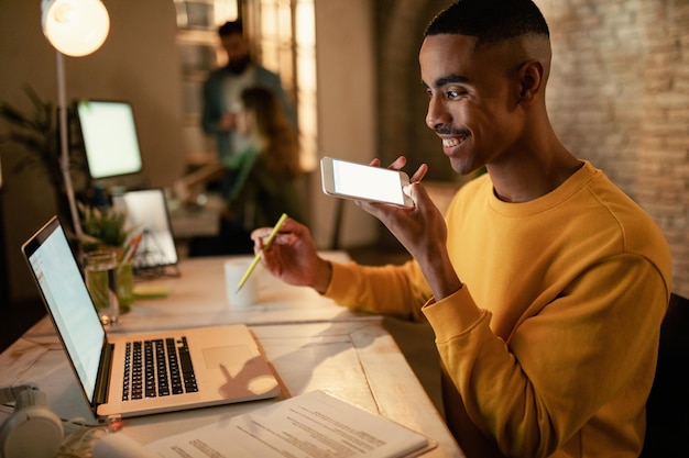 Happy black entrepreneur communicating over mobile phone's speaker while using computer and working in the evening at his office