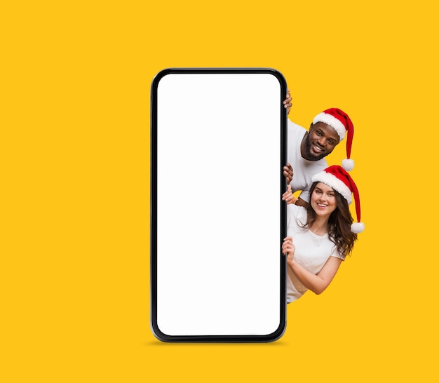 Happy black and european young couple in Santa Claus hats peeking out from big smartphone with empty screen