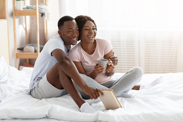 Happy black family staying home on weekend, sitting on bed, drinking tea and reading book, looking at copy space. Cheerful african american couple enjoying time together, spending day in bed