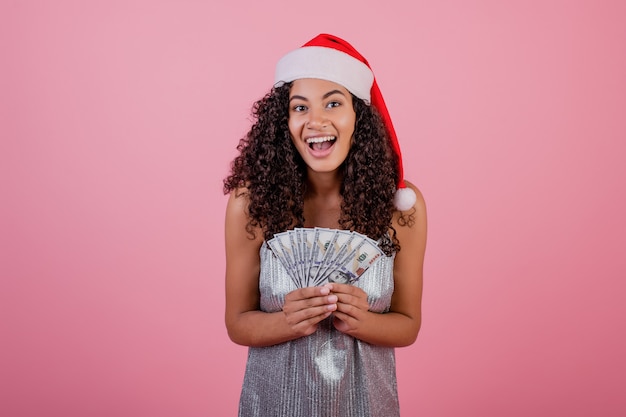 Happy black girl holding one hundred dollar bills wearing santa hat isolated