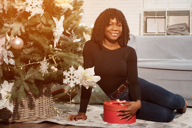 Happy black girl with curly hair sits near a Christmas tree and holds a gift, looking at camera